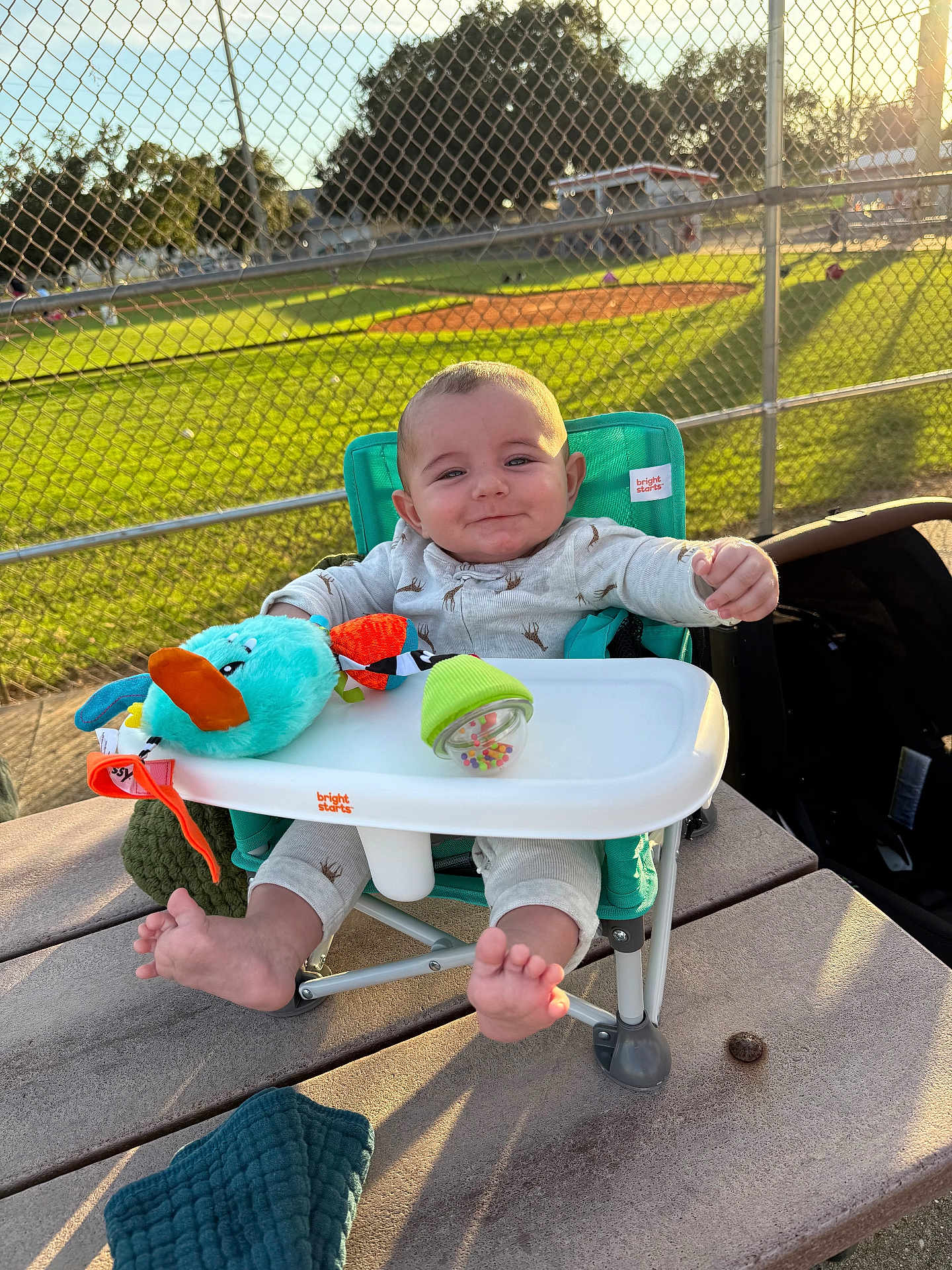 Gianni is registered to the contest to win money with this photo: baby, high_chair, toy, outdoor, sunlight, park, fence, baseball_field, green_grass, smiling, child, onesie, feet, table, playful, daylight, person, seat, happy, infant