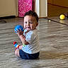 toddler, child, baby, indoor, floor, ball, toy, colorful_socks, white_shirt, navy_shorts, curly_hair, smiling, sitting, playful, wooden_floor, pink_curtain, room, casual_clothing, happy, person