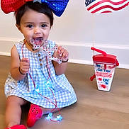 Eliza is registered to the contest to win money with this photo: toddler, child, bow, red_shoes, blue_dress, toy, star_shaped, necklace, glowing_lights, wooden_floor, patriotic, american_flag, drink_cup, indoor, sitting, cute, playful, festive, smiling, happy