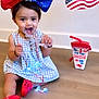 toddler, child, bow, red_shoes, blue_dress, toy, star_shaped, necklace, glowing_lights, wooden_floor, patriotic, american_flag, drink_cup, indoor, sitting, cute, playful, festive, smiling, happy