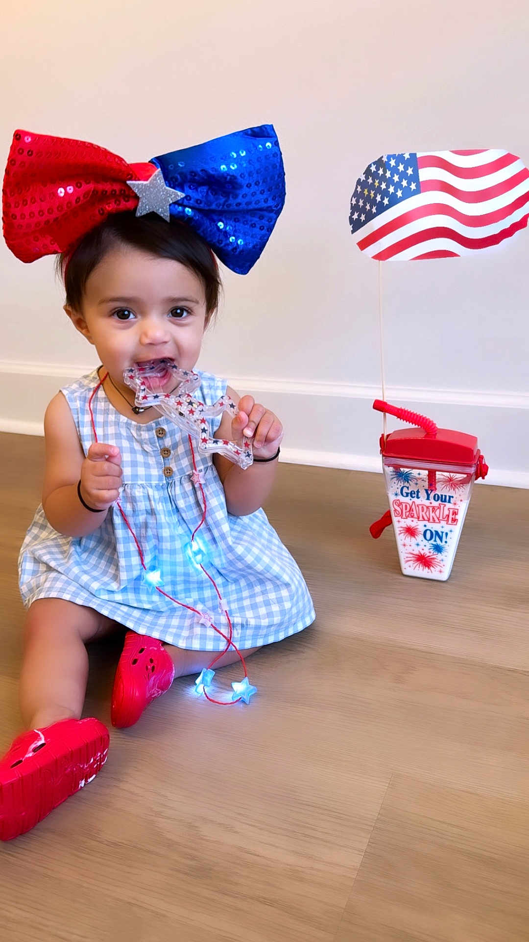 Eliza is registered to the contest to win money with this photo: toddler, child, bow, red_shoes, blue_dress, toy, star_shaped, necklace, glowing_lights, wooden_floor, patriotic, american_flag, drink_cup, indoor, sitting, cute, playful, festive, smiling, happy