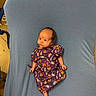 baby, infant, purple_gown, patterned_clothing, blue_blanket, chair, indoor, newborn, cute, small, lying_down, curious, human, child, soft_texture, fabric, colorful, portrait, face, skin