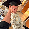 Sonny is registered to the contest to win money with this photo: baby, infant, child, graduation_cap, onesie, hand, indoor, person, expression, cute, newborn, clothing, holding, surprised, face, headwear, room, background, adult_hand, human