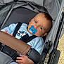 baby, pacifier, stroller, child, infant, blue_clothing, gray_fabric, brown_handle, straps, outdoor, toddler, cute, person, legs, hand, seat, wheel, socks, face, head