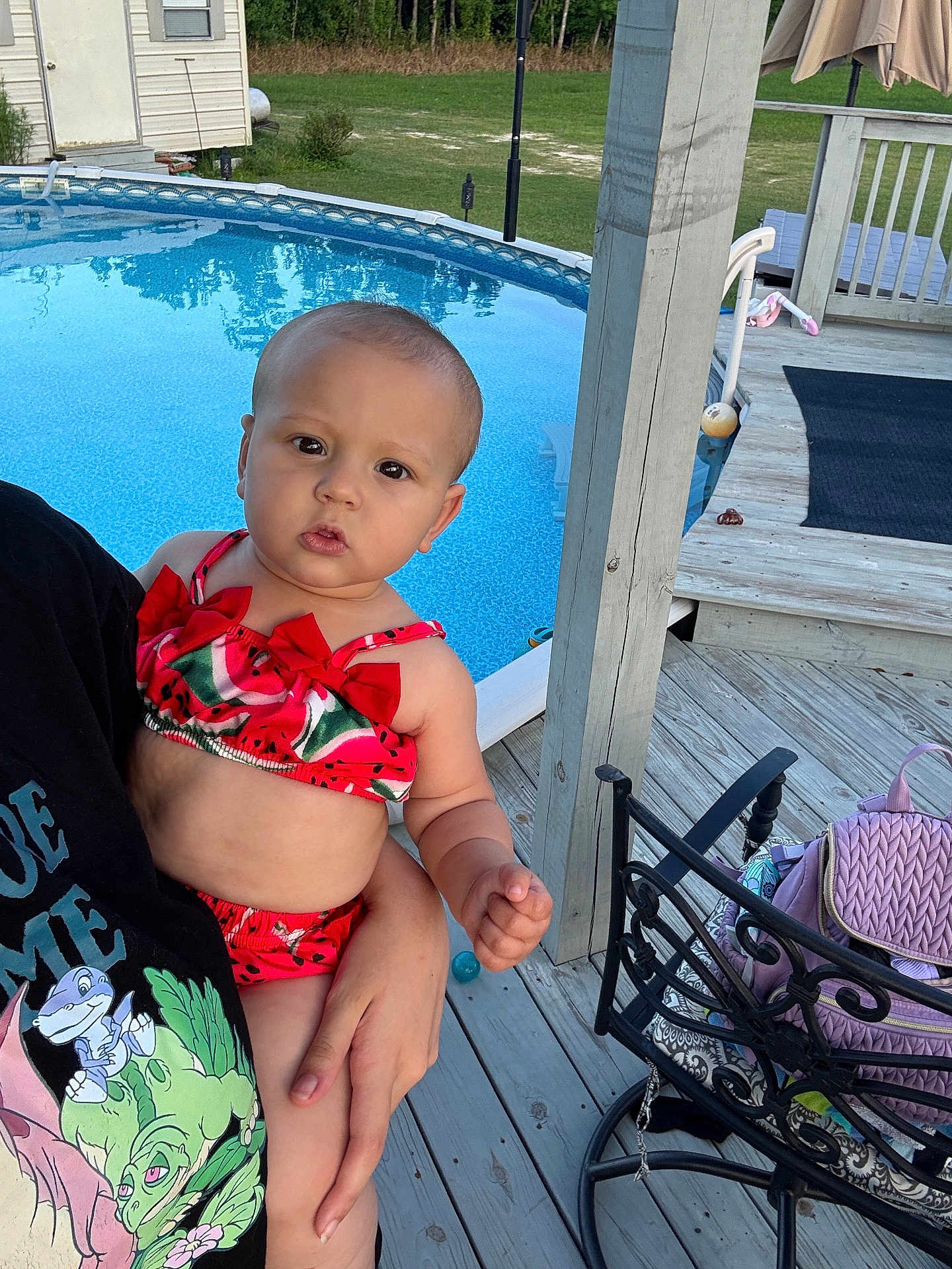 Nyana is registered to the contest to win money with this photo: baby, child, swimsuit, watermelon, pool, outdoor, deck, wood, hand, person, clothing, furniture, chair, backyard, summer, daylight, expression, curious, holding, portrait