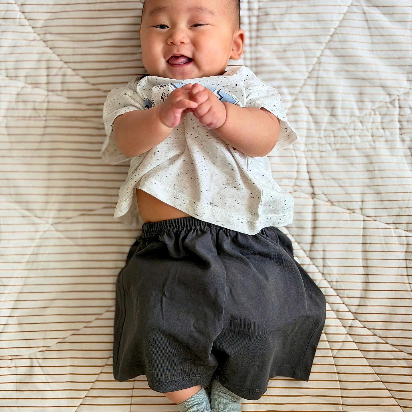 Evan joined the competition — help win amazing prizes! baby, black_shorts, cheerful, child, clapping, cute, fabric, feet, hands, happy, indoors, infant, lying_down, playful, portrait, smiling, socks, striped_quilt, white_shirt, young_child