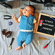 Evan joined the competition — help win amazing prizes! baby, blue_outfit, child, cute, face, foot, hand, indoor, infant, lying_down, measuring_tape, person, portrait, screwdrivers, signboard, striped_sheet, text, tools, toy_car, wrenches
