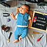 baby, blue_outfit, child, cute, face, foot, hand, indoor, infant, lying_down, measuring_tape, person, portrait, screwdrivers, signboard, striped_sheet, text, tools, toy_car, wrenches