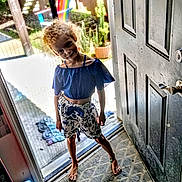 Stella joined the competition — help win amazing prizes! child, barefoot, doorway, blue_top, floral_shorts, curly_hair, indoor, outdoor, potted_plants, colorful_chair, playful, smiling, shadow, door, floor_mat, sunlight, plants, wooden_fence, casual_clothing, standing