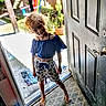 barefoot, blue_top, casual_clothing, child, colorful_chair, curly_hair, door, doorway, floor_mat, floral_shorts, indoor, outdoor, plants, playful, potted_plants, shadow, smiling, standing, sunlight, wooden_fence
