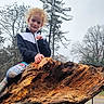 bare_trees, casual_clothing, child, curly_hair, daytime, fall, grass, jacket, leggings, leisure, nature, outdoor, overcast_sky, park, person, playful, smiling, tree_trunk, wood, young_person