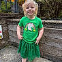 child, girl, toddler, unicorn_shirt, green_shirt, green_skirt, pigtails, smile, sandals, outdoor, stone_wall, railing, concrete_ground, bushes, plants, porch_steps, portrait, person, happy, casual_clothing