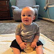 James joined the competition — help win amazing prizes! baby, child, infant, smiling, sitting, blanket, clothing, nike, shorts, tshirt, wooden_sign, carpet, chair, dresser, room, home_interior, face, hands, feet, happy