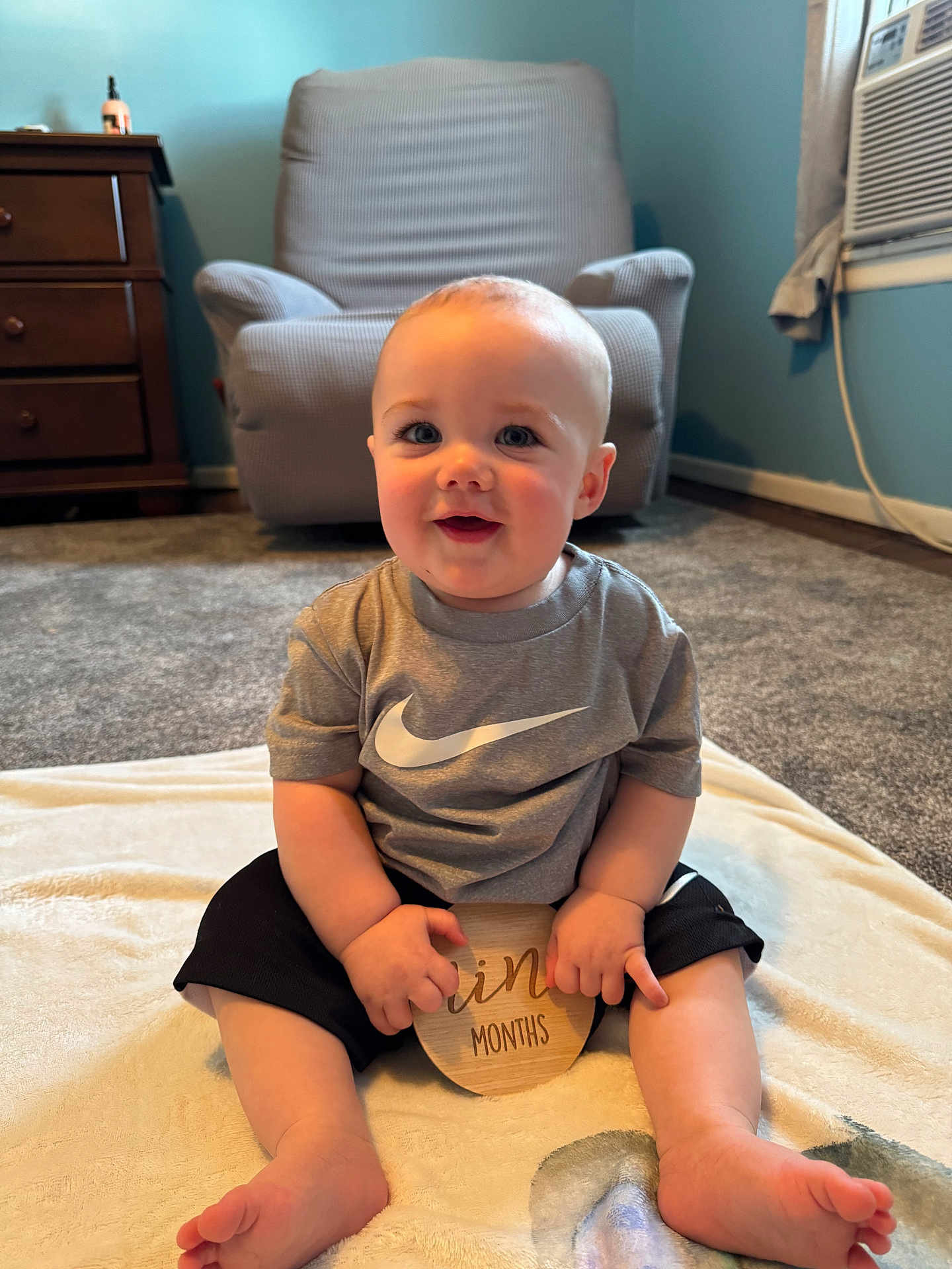 James joined the competition — help win amazing prizes! baby, child, infant, smiling, sitting, blanket, clothing, nike, shorts, tshirt, wooden_sign, carpet, chair, dresser, room, home_interior, face, hands, feet, happy