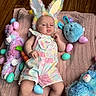 Riley is registered to the contest to win money with this photo: baby, bunny_ears, stuffed_animals, easter_eggs, pink_blanket, wood_floor, colorful_clothing, pastel_colors, toy_carrot, blue_toy, purple_toy, baby_onesie, infant, lying_down, indoor, cute, child, holiday_theme, soft_toys, playful