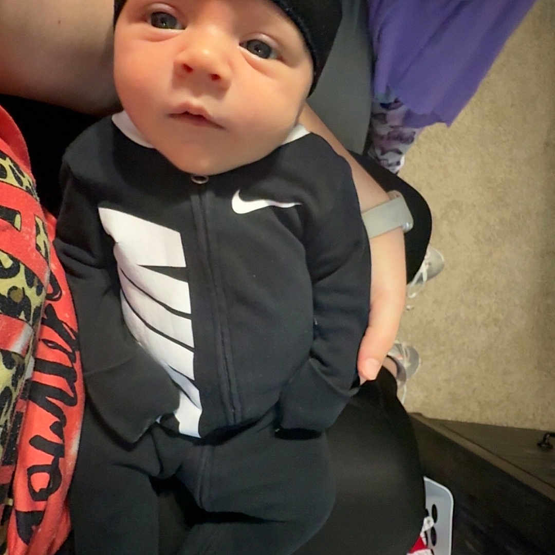 Hunner joined the competition — help win amazing prizes! baby, infant, black_hat, black_outfit, nike_logo, person, hand, laundry_basket, carpet, purple_clothing, sitting, indoor, curious_expression, footwear, child, home, casual, portrait, young_child, person_holding_baby