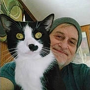 Rascal joined the competition — help win amazing prizes! cat, person, indoor, smiling, beanie, black_and_white_cat, yellow_eyes, portrait, wooden_wall, framed_art, closeup, whiskers, happy, feline, casual_clothing, home, pet, companion, face, cozy