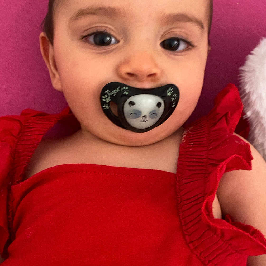Inaya a rejoint le concours — aidez-le/la à gagner de superbes lots ! adorable, baby, child, close_up, clothing, cute, expression, eyes, face, head, indoor, infant, pacifier, person, pink_background, portrait, red_dress, ruffles, skin, young