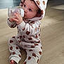baby, child, bottle, onesie, hood, flooring, indoor, infant, cute, drinking, sitting, cozy, curly_hair, patterned_clothing, soft_texture, footwear_off, portrait, young_child, warm_clothing, home