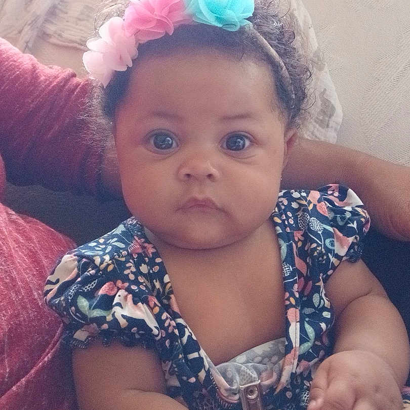 Za'Karrah joined the competition — help win amazing prizes! arm, baby, blue_eyes, child, closeup, clothing, couch, curly_hair, cute, expression, flower_headband, hand, indoor, infant, patterned_dress, person, portrait, sitting, skin, soft_light