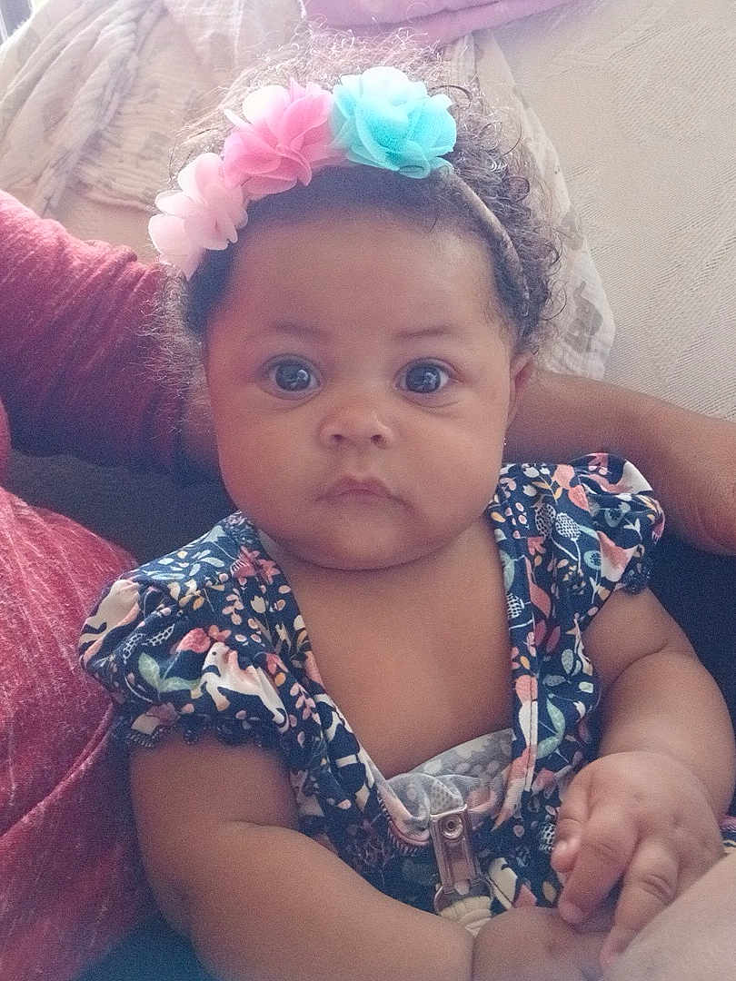 Za'Karrah joined the competition — help win amazing prizes! baby, child, flower_headband, curly_hair, blue_eyes, sitting, person, arm, clothing, patterned_dress, indoor, couch, soft_light, closeup, portrait, cute, infant, skin, hand, expression