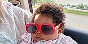Za'Karrah is registered to the contest to win money with this photo: baby, child, sunglasses, car_interior, curly_hair, onesie, animal_pattern, person, hand, window, daylight, cute, serious_expression, infant, clothing, holding, portrait, indoor, car_seat, relaxing