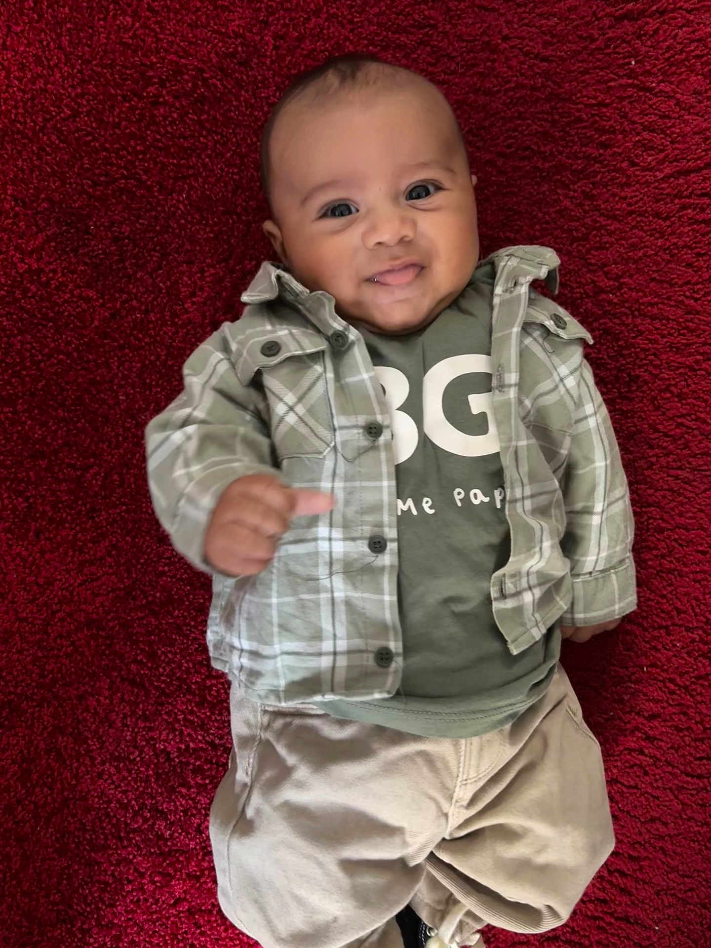 Léandro participe au concours pour gagner de l'argent avec cette photo : baby, infant, smiling, lying_down, red_rug, plaid_shirt, green_tshirt, pants, portrait, face, eyes, hand, skin, clothing, cute, adorable, brown_eyes, casual_outfit, indoor, closeup