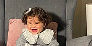 Eline a rejoint le concours — aidez-le/la à gagner de superbes lots ! toddler, child, smiling, curly_hair, bow, gray_armchair, pink_pillow, plant, white_collar, gray_sweater, white_tights, happy, indoor, portrait, sitting, cozy, cute, baby, furniture, home