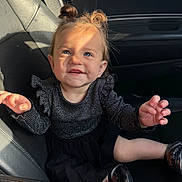 Melody is registered to the contest to win money with this photo: child, toddler, car_seat, black_dress, black_shoes, hair_buns, smiling, blue_eyes, sunlight, indoor, seat, person, cute, happy, portrait, baby, clothing, hands, face, car_interior
