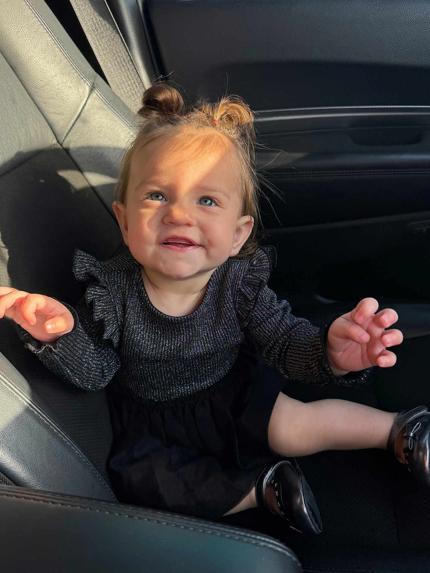 Melody is registered to the contest to win money with this photo: child, toddler, car_seat, black_dress, black_shoes, hair_buns, smiling, blue_eyes, sunlight, indoor, seat, person, cute, happy, portrait, baby, clothing, hands, face, car_interior