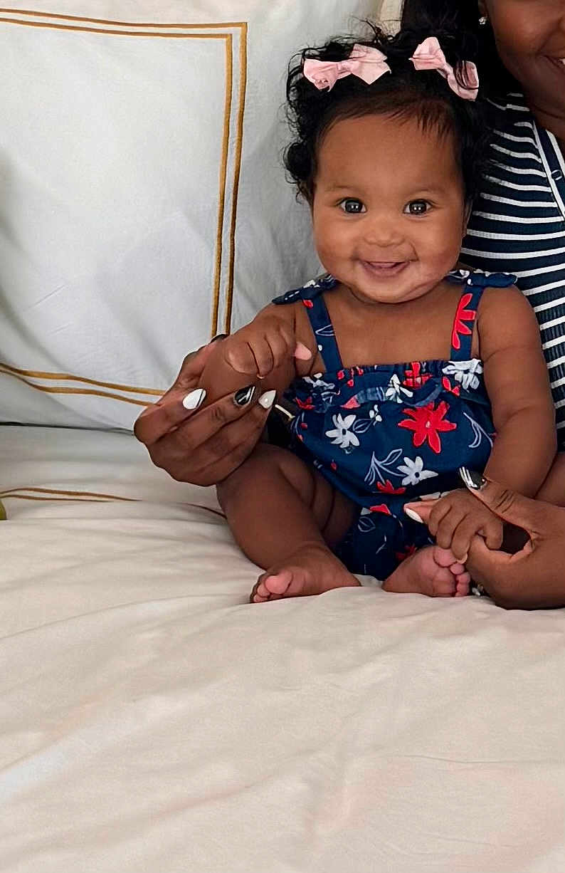 Elizah is registered to the contest to win money with this photo: baby, child, smiling, curly_hair, pink_bows, blue_flowered_dress, hands, adult_hands, nail_art, bed, white_bedspread, striped_clothing, person, indoor, happy, cute, infant, holding_hands, portrait, family