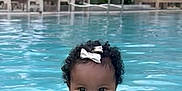 Elizah joined the competition — help win amazing prizes! baby, child, swimming_pool, water, smiling, curly_hair, bow, swimsuit, outdoor, summer, happy, person, poolside, sunlight, relaxation, leisure, vacation, lounge_chair, greenery, daytime