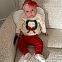 baby, infant, child, sitting, couch, sweater, red_pants, headband, bow, wreath, white_socks, indoors, portrait, looking_at_camera, curious, soft_light, beige_couch, knitwear, holiday_theme, cozy
