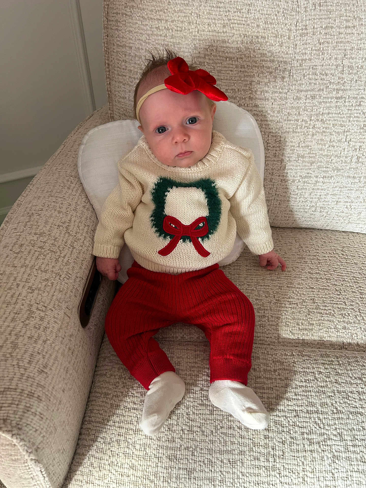 Capri joined the competition — help win amazing prizes! baby, infant, child, sitting, couch, sweater, red_pants, headband, bow, wreath, white_socks, indoors, portrait, looking_at_camera, curious, soft_light, beige_couch, knitwear, holiday_theme, cozy