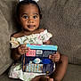 barefoot, book, brown_skin, child, clothing, couch, curly_hair, cute, dress, expression, furniture, holding, indoors, looking, person, portrait, sitting, toddler, white_bow, young
