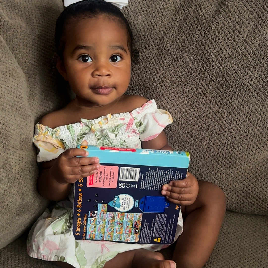 Aiyara is registered to the contest to win money with this photo: barefoot, book, brown_skin, child, clothing, couch, curly_hair, cute, dress, expression, furniture, holding, indoors, looking, person, portrait, sitting, toddler, white_bow, young