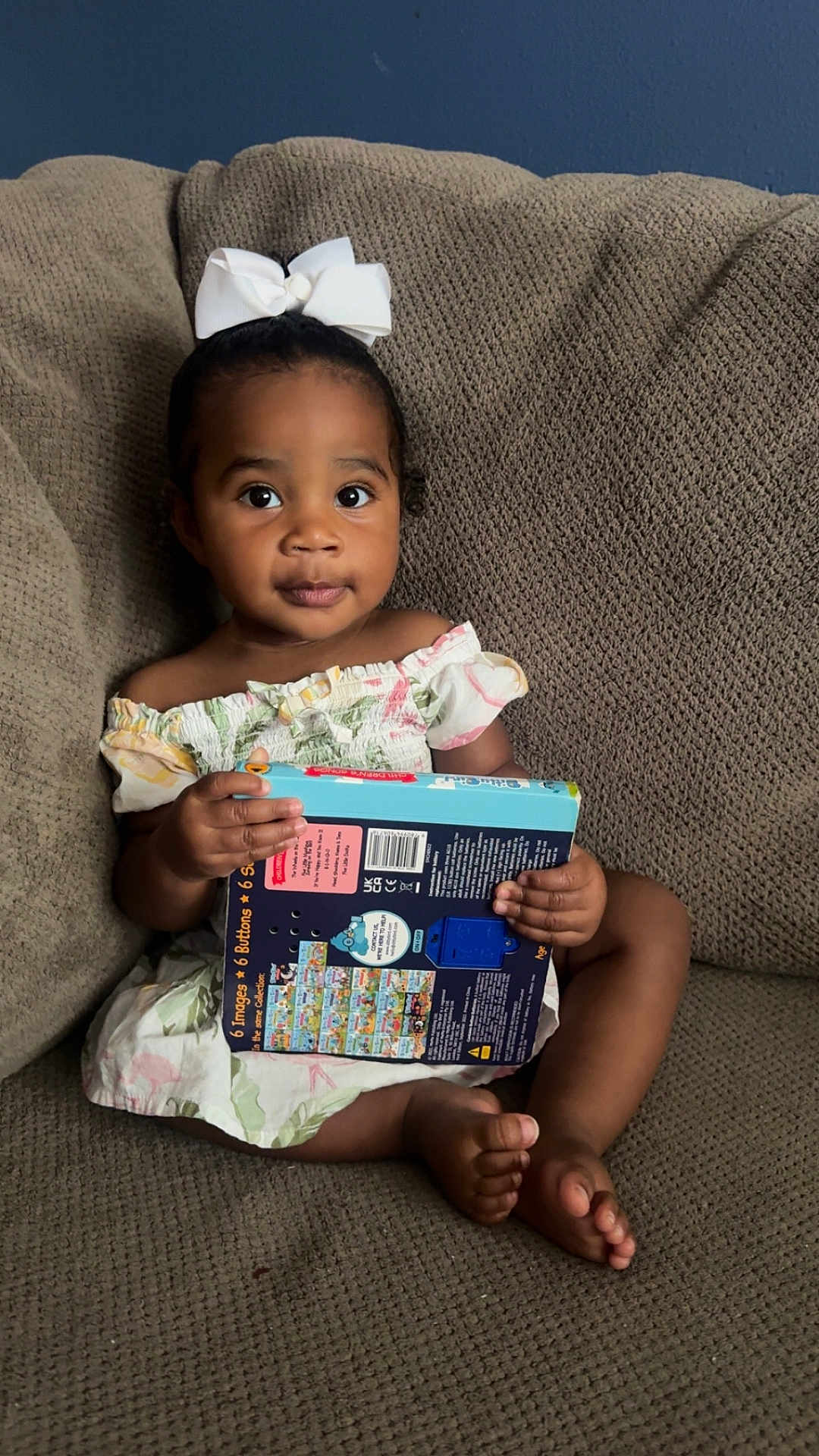 Aiyara is registered to the contest to win money with this photo: barefoot, book, brown_skin, child, clothing, couch, curly_hair, cute, dress, expression, furniture, holding, indoors, looking, person, portrait, sitting, toddler, white_bow, young