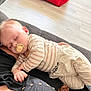 baby, child, sleeping, pacifier, striped_shirt, pants, couch, hand, indoor, floor, red_pillow, black_clothing, nap_time, cozy, person, relaxing, comfort, home, rest, casual