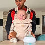 baby, adult, baby_carrier, smiling, indoor, table, baby_bottle, person, clothing, happy, face, modern_interior, parent, holding, head, sitting, brown_pants, striped_shirt, short_hair, facial_expression