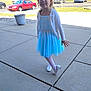 Addie is registered to the contest to win money with this photo: child, girl, blue_dress, bow, white_cardigan, outdoor, sidewalk, pose, smile, curly_hair, sunlight, cars, parking_lot, concrete, footwear, white_shoes, playful, daytime, nature, grass