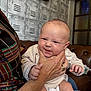 baby, infant, hand, person, adult, face, expression, indoor, clothing, sitting, couch, furniture, skin, head, holding, wrinkle, smile, scrunched_face, background, pattern