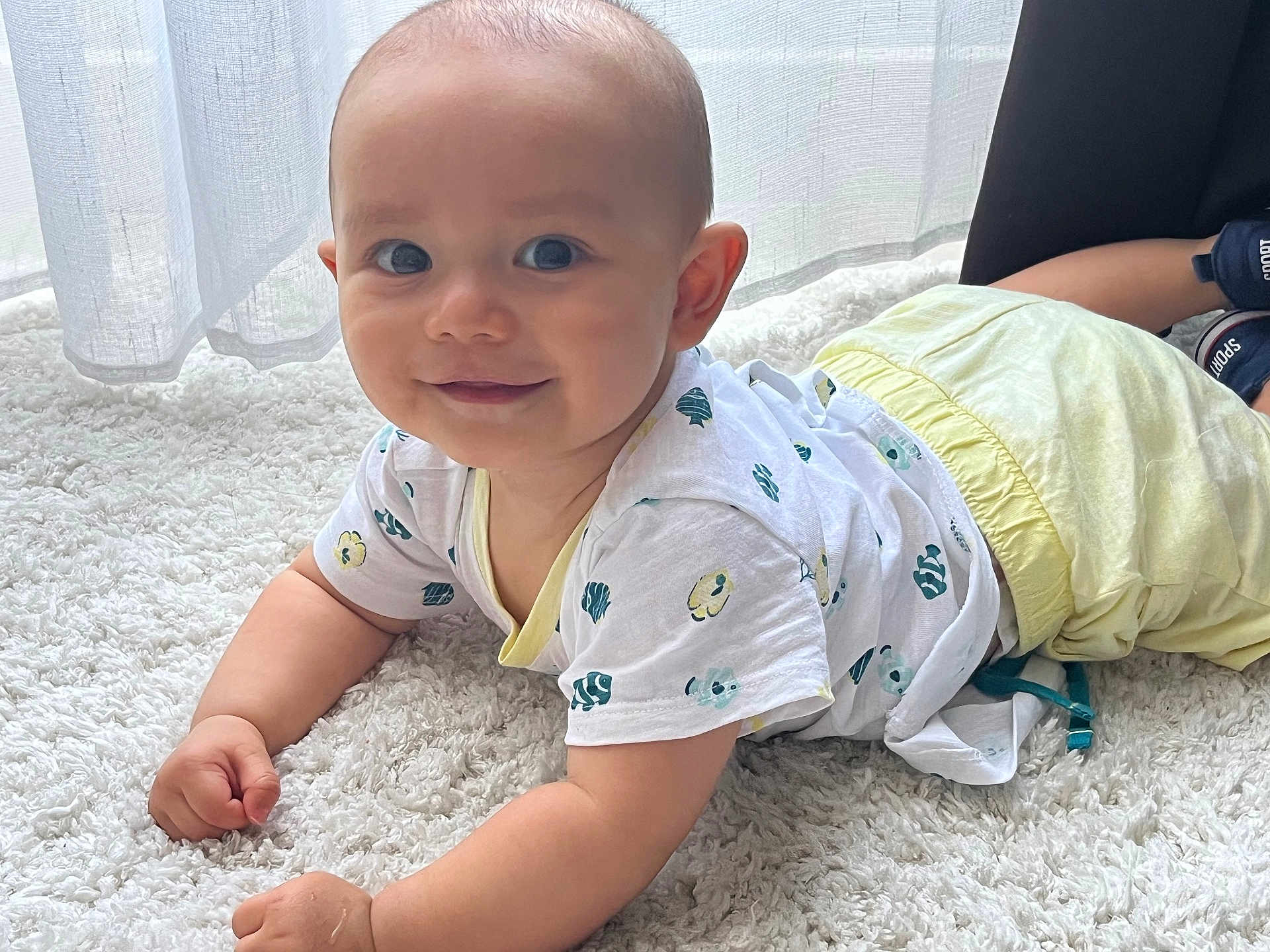 Léandre participe au concours pour gagner de l'argent avec cette photo : arms, baby, carpet, child, clothing, crawling, curtains, face, floor, happy, indoor, infant, natural_light, pants, patterned_clothing, person, smile, tshirt, white_carpet, window