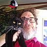 beard, black_cat, casual, cat, cozy, facial_hair, feline, glasses, hat, indoor, man, pet, pipe, plant, portrait, red_shirt, selfie, smile, whiskers, yellow_eyes