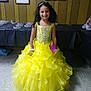 child, girl, yellow_dress, curly_hair, smiling, tiaras, pageant, dress, party, event, floor, portrait, indoor, celebration, person, jewelry, bracelet, black_tablecloth, wood_paneling, decoration