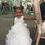 child, girl, smiling, white_dress, formal_wear, dress_shop, mirror, carpet, chair, indoor, happy, person, clothing, hairstyle, headband, reflection, store, retail, fashion, young