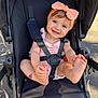 baby, infant, stroller, headband, bow, smile, toes, hands, safety_harness, seat, outdoor, sunlight, cute, pink_clothing, happy, portrait, pavement, leaf, child, playful