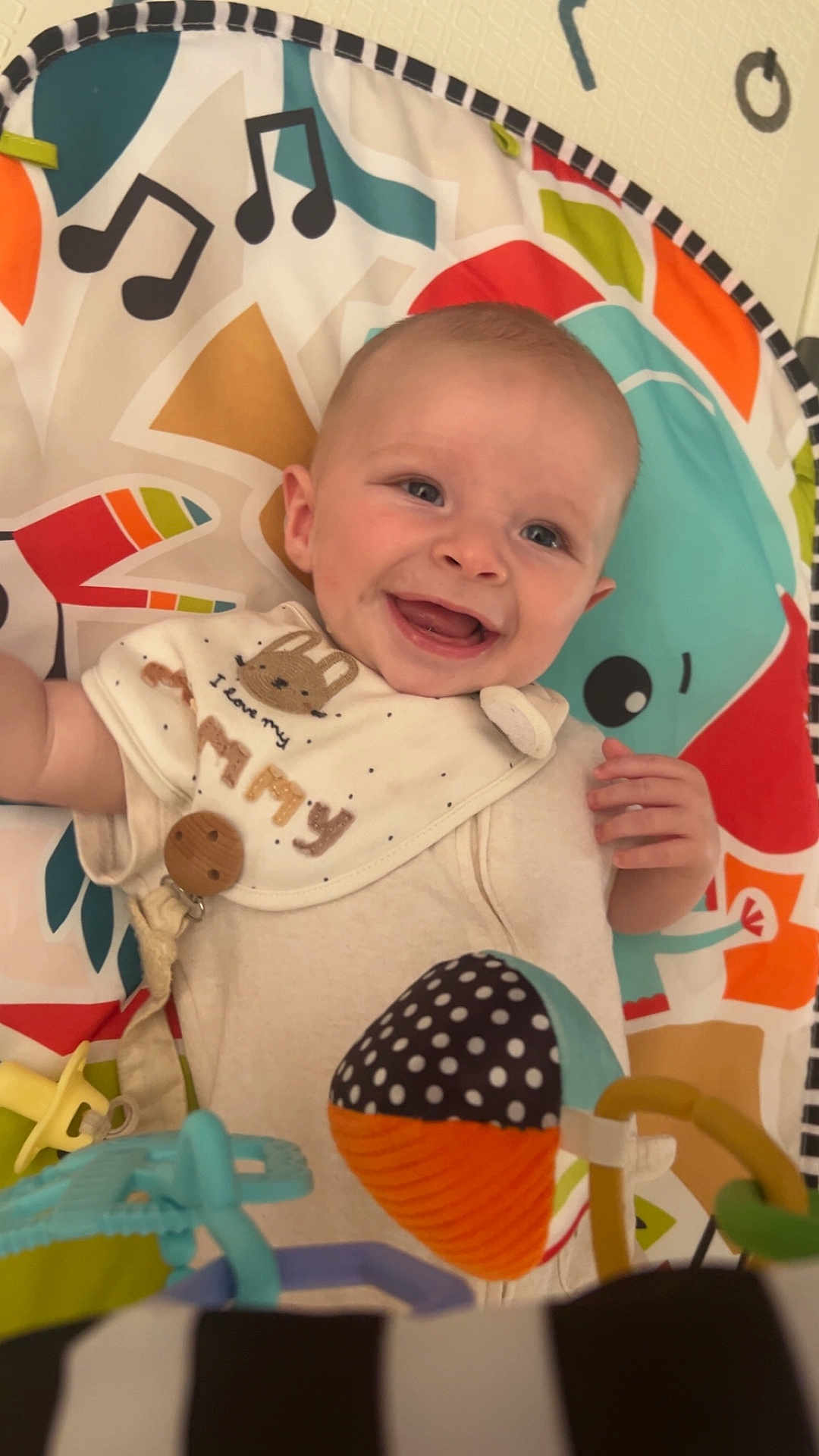 Archie is registered to the contest to win money with this photo: baby, smiling, play_mat, colorful, bib, toy, infant, happy, face, child, cute, indoor, closeup, polka_dot, music_notes, hand, blue_eyes, fabric, pattern, soft_toys
