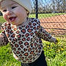 toddler, child, smiling, beanie, sweater, heart_pattern, outdoor, grass, dandelion, fence, sunny, park, happy, nature, playful, cute, person, young_child, casual_clothing, daytime