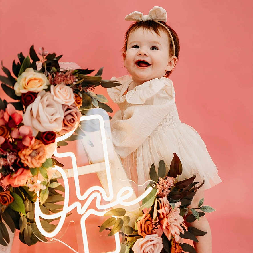 Oaklynne joined the competition — help win amazing prizes! baby, child, girl, smiling, barefoot, pink_background, flower_arrangement, neon_sign, first_birthday, headband, dress, happy, portrait, studio_photo, decor, celebration, cute, one_year_old, standing, holding