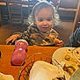 toddler, child, curly_hair, camouflage_shirt, restaurant, table, bread, sippy_cup, plate, food, napkin, basket, chair, window, indoor, person, eating, meal, happy, casual